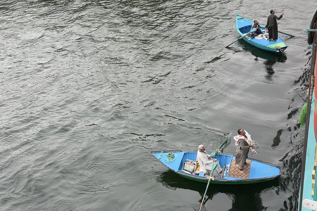 Galabeya sellers in two rowing boats beside a Nile cruise ship at Esna, one holding up cloth to the deck